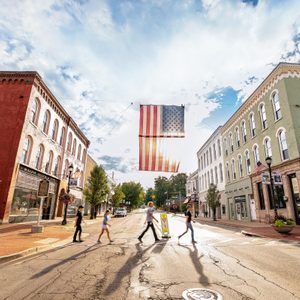 People walk across a street beneath a large American flag in a small town, surrounded by historical brick buildings under a cloudy sky.