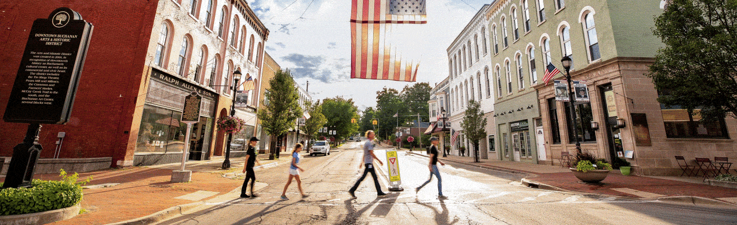 Four people walk across a crosswalk; a large American flag hangs above a quaint downtown street lined with historic buildings.