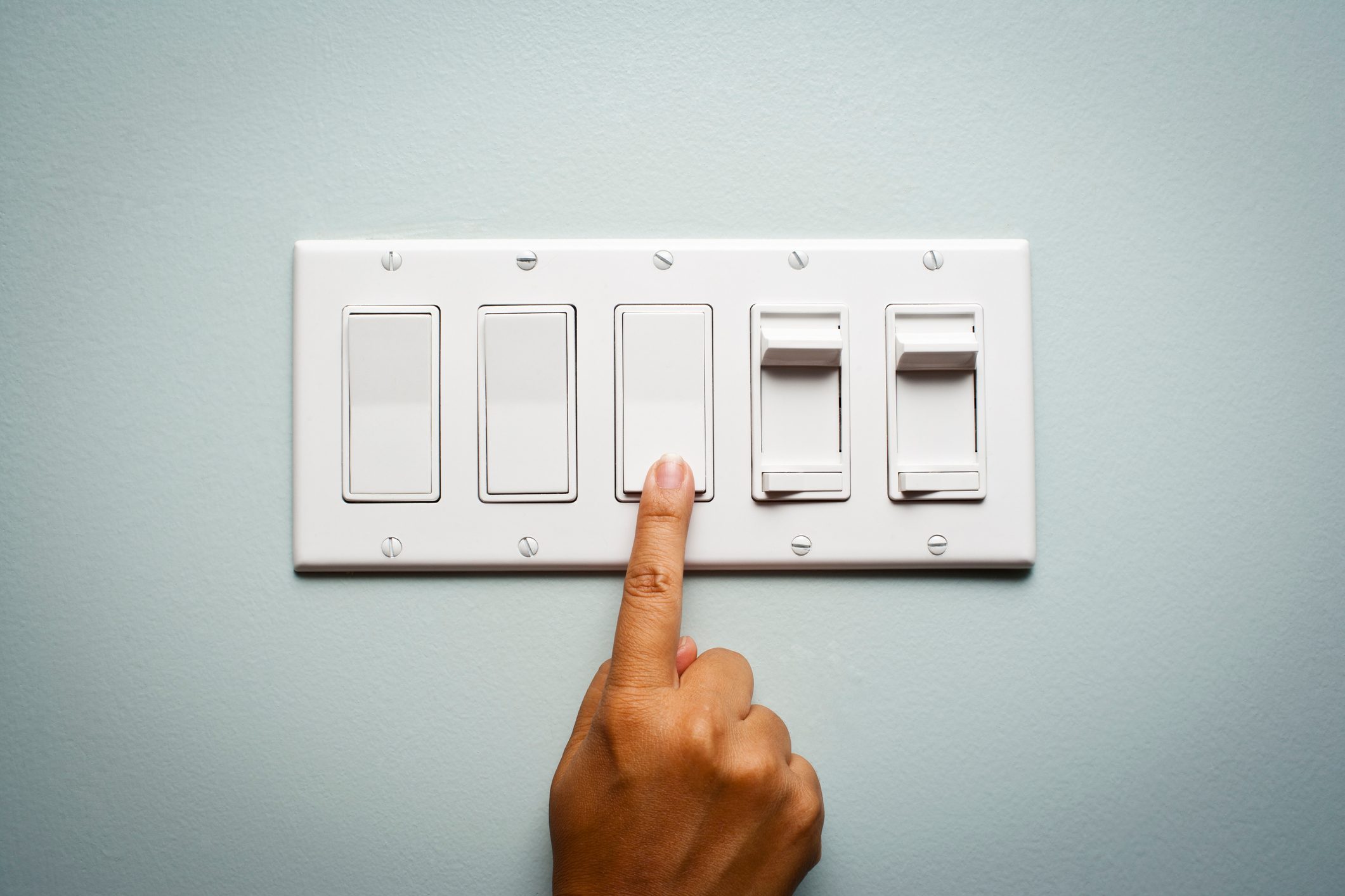 A hand presses a white light switch on a pale blue wall with three additional switches.