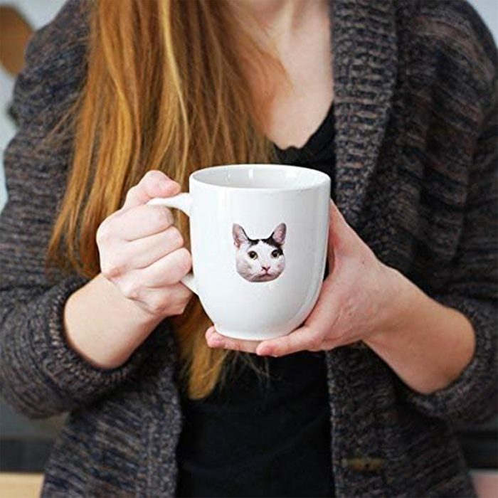 Hands hold a white mug showing a cat face, worn sweater visible, indoors.