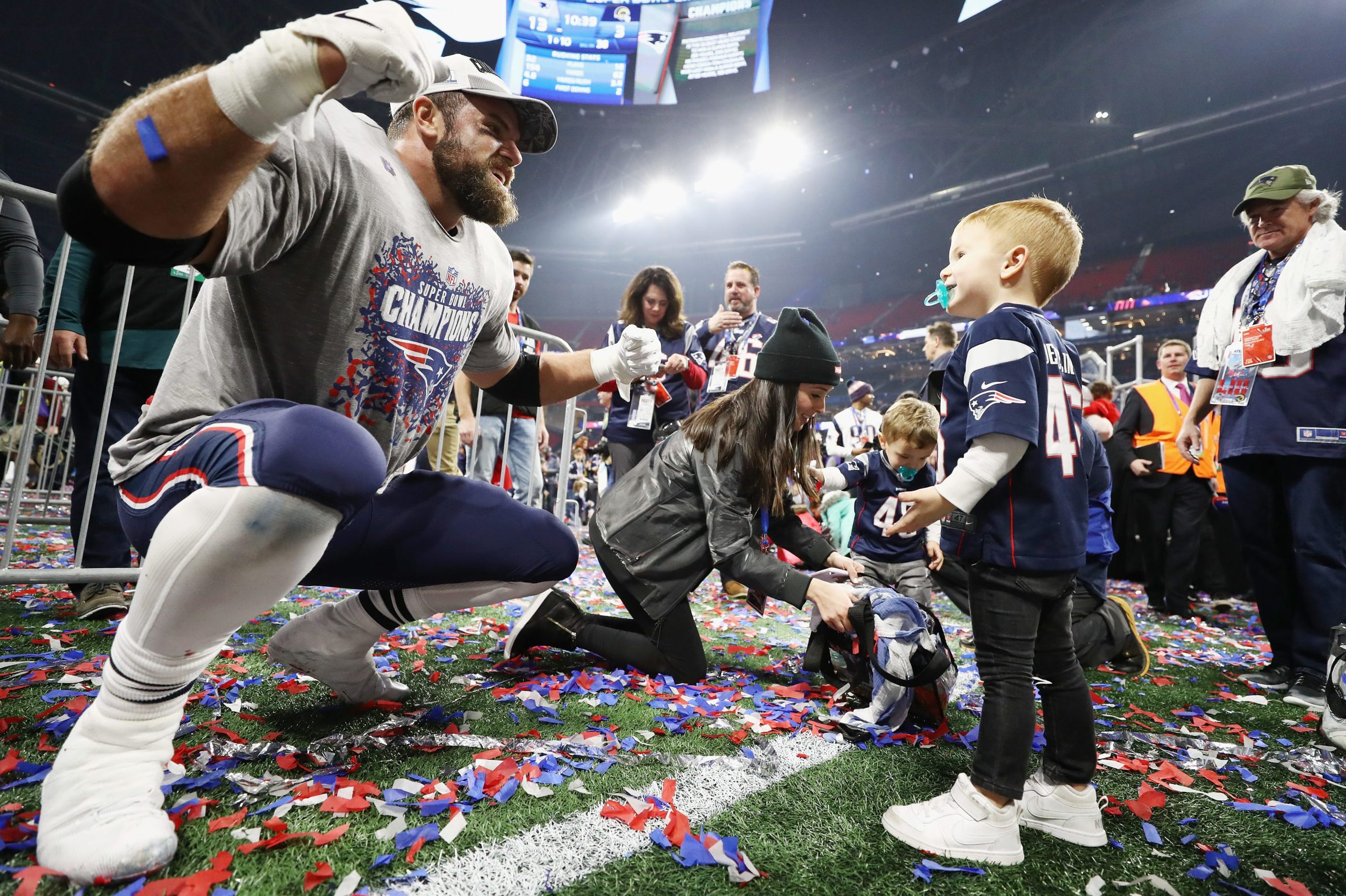 Football player celebrating on confetti-covered field, interacting with young fan wearing a jersey. Stadium lights and crowd in the background.