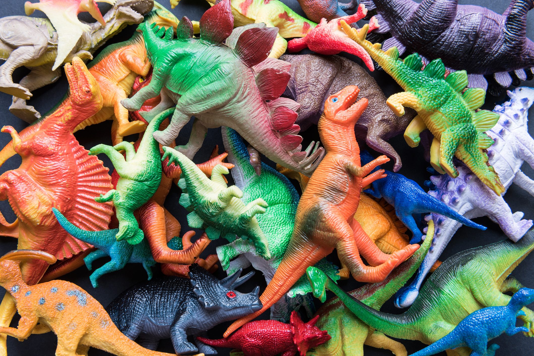 Toy dinosaurs scattered randomly in a colorful pile against a dark background, displaying various shapes and bright colors.