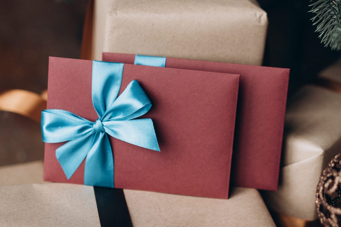 Red envelopes with blue ribbons rest against wrapped gifts, placed in a festive setting with soft lighting and evergreen branches nearby.