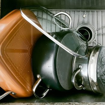 Pots and a glass lid stack in a stainless steel sink, draining water, on a granite countertop background.