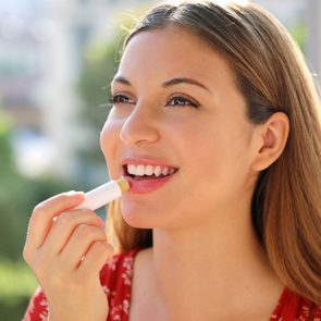A person applies lip balm in an outdoor setting with blurred green foliage in the background.