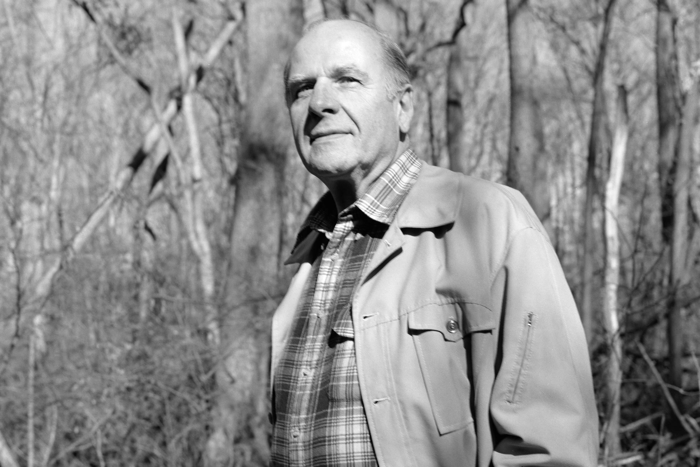 Man stands calmly in a forest, wearing a plaid shirt and light jacket. The trees and branches appear bare, suggesting a winter setting.