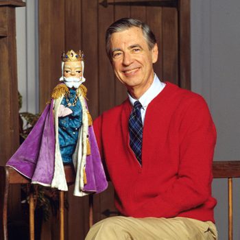 A man in a red sweater smiles beside a puppet king wearing a purple robe in a cozy, wooden interior setting.