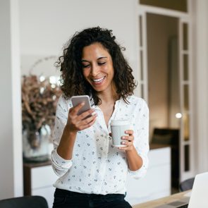 A person smiles at a phone, holding a coffee cup, seated in a bright room with a laptop and decor.