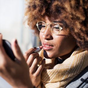 A person applies lipstick, looking into a small mirror, wearing glasses and a scarf, in a brightly lit room.