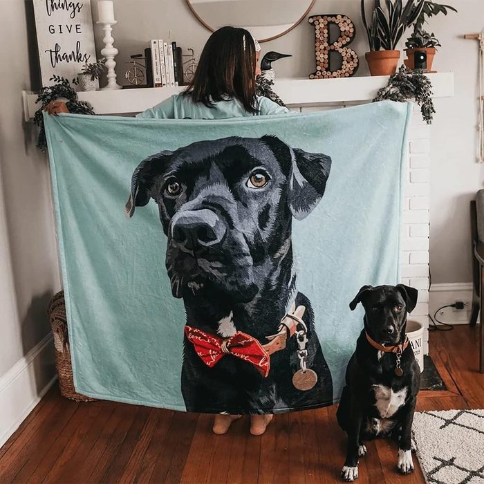 Person holding blanket depicting a black dog with red bow tie; actual black dog sits beside, indoors with decorated mantel.