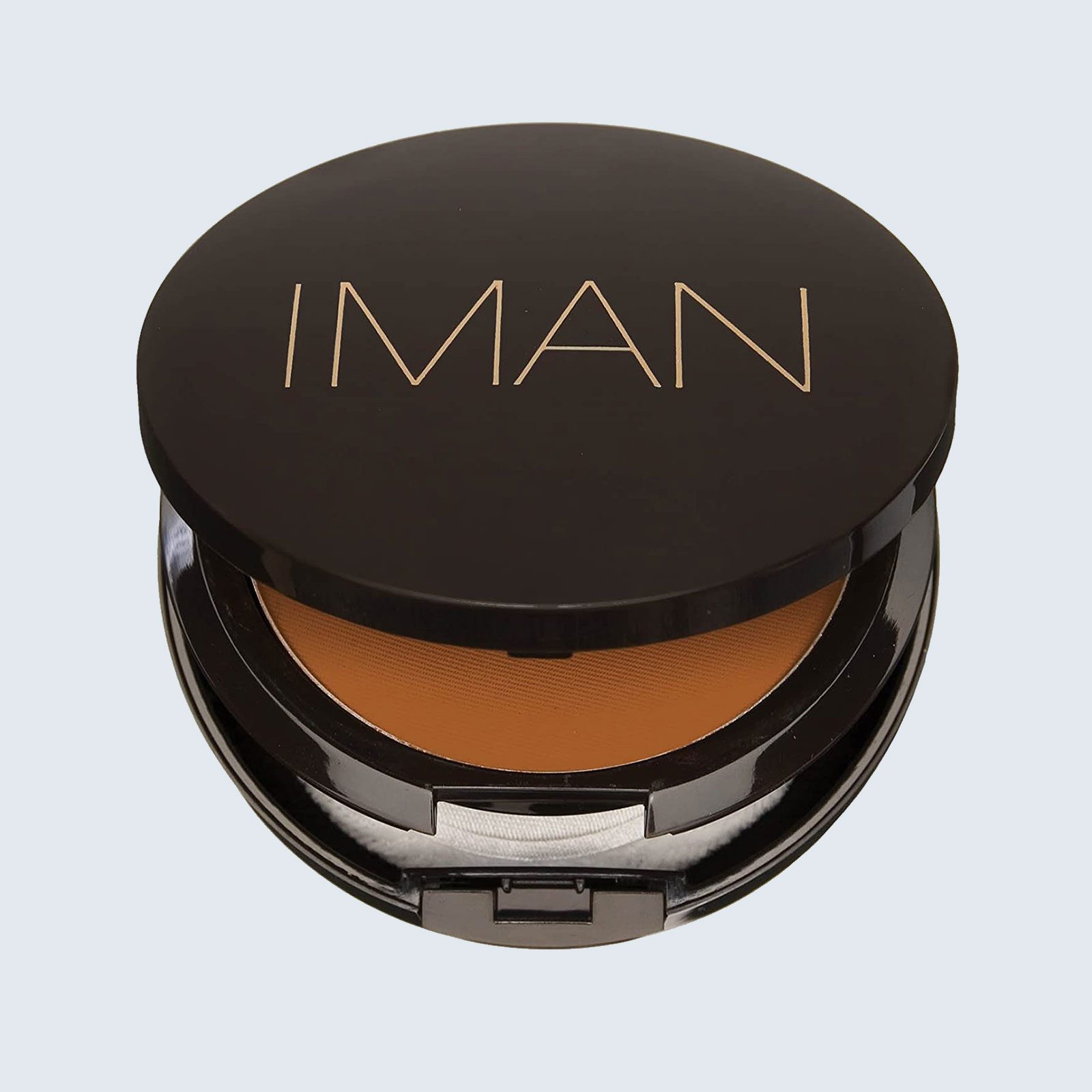 Compact makeup case opens slightly, revealing brown powder and applicator, against a simple light background. Text on lid reads "IMAN."