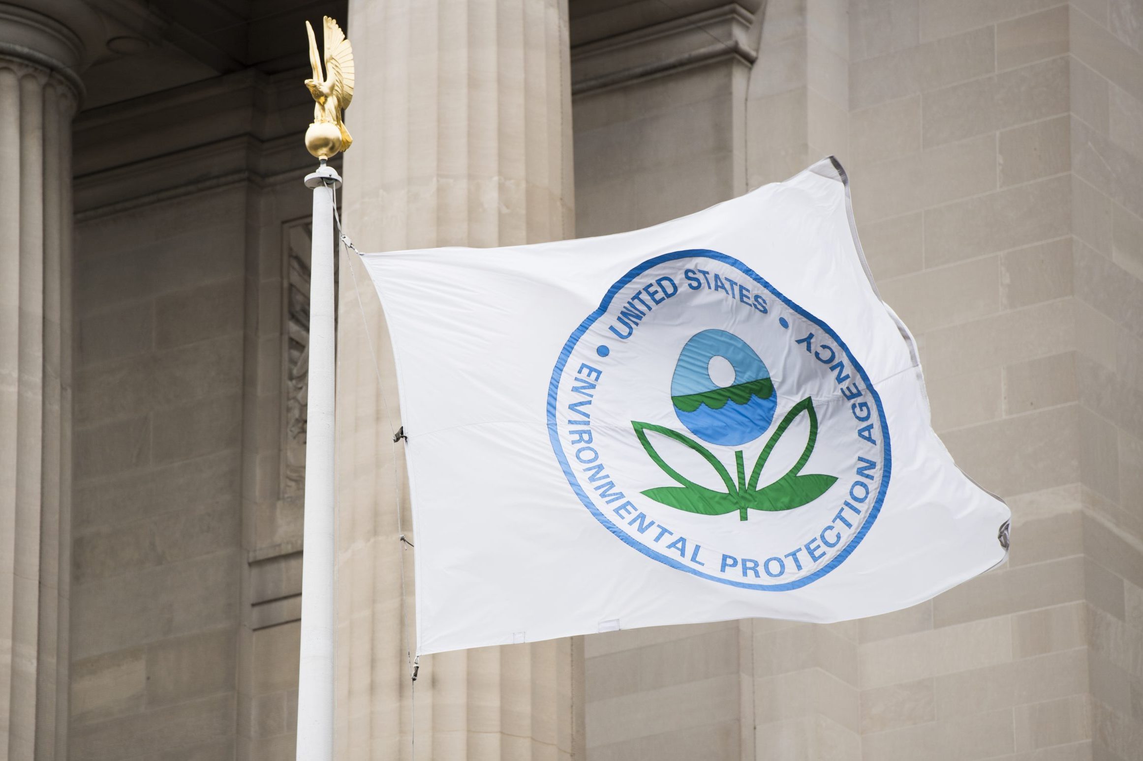 Flag with Environmental Protection Agency logo waves on a pole, next to a stone building with columns.