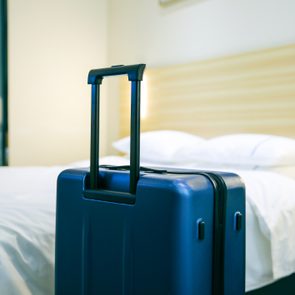 A blue suitcase stands upright beside a neatly made bed in a hotel room.