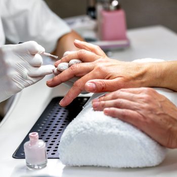 Hands paint fingernails on a womanâ€™s hand using a brush, while resting on a white towel in a nail salon.