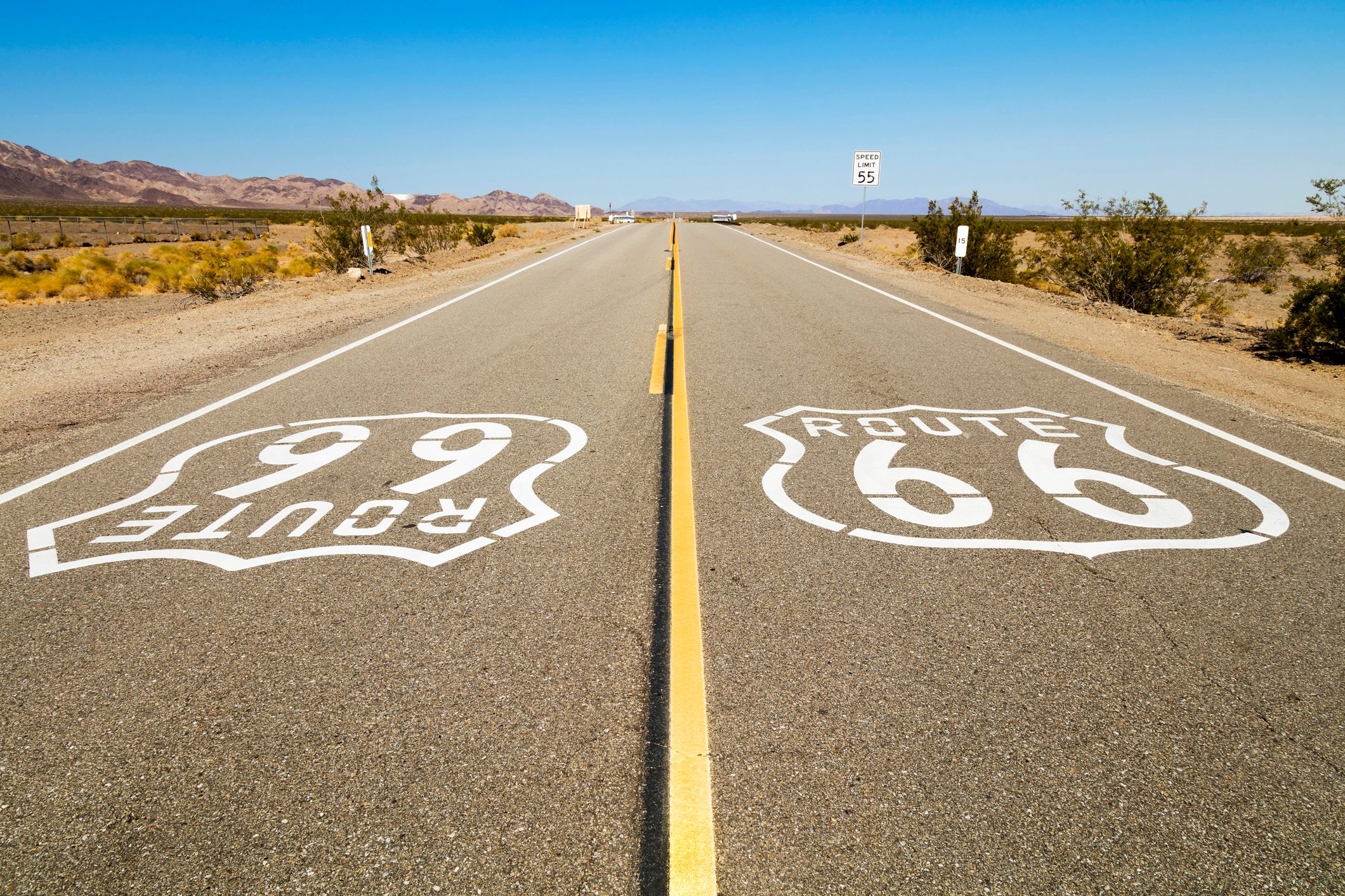 Route 66 marked on empty desert road, yellow center line, bushes, and distant mountains under clear blue sky. Speed sign shows "SPEED LIMIT 55".