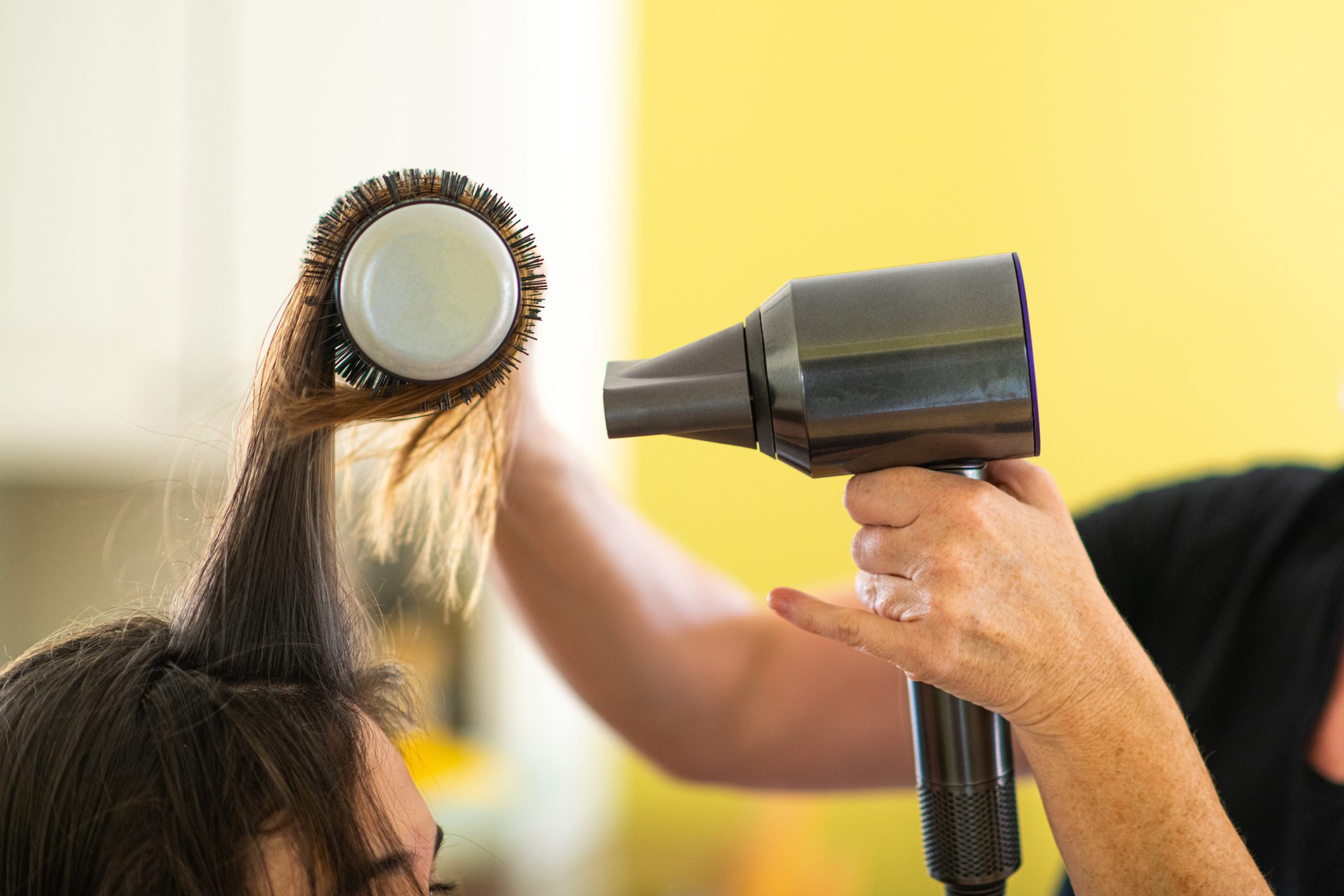 A round brush holds hair while a person uses a hairdryer in a brightly lit room with a yellow wall.