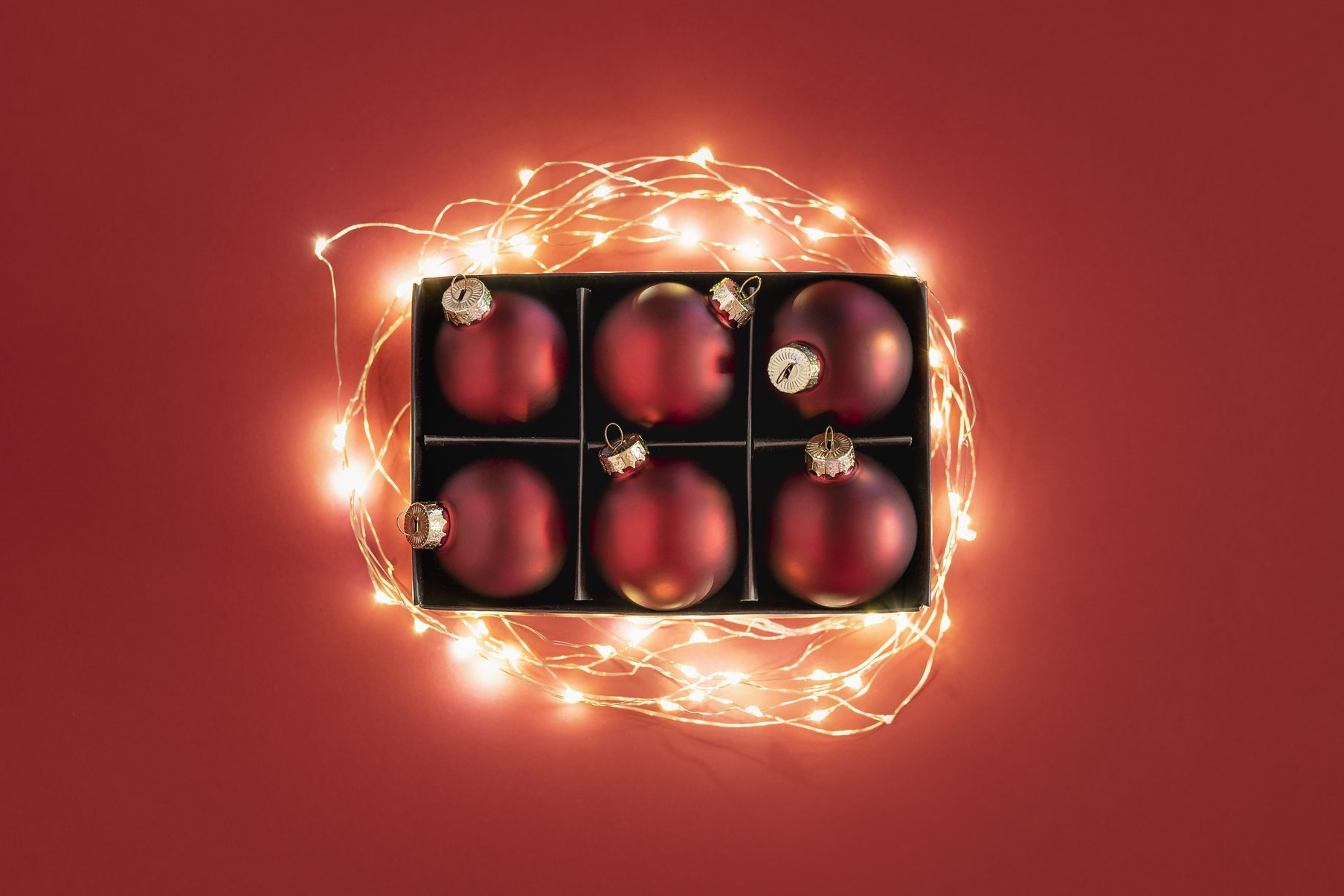 Ornaments rest in a box, surrounded by glowing fairy lights on a solid red background.