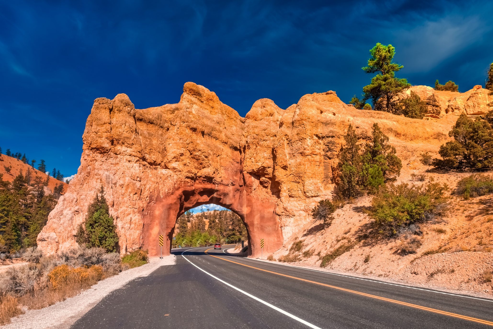 Road curves through a natural rock tunnel, surrounded by red rock formations and sparse vegetation under a clear blue sky.