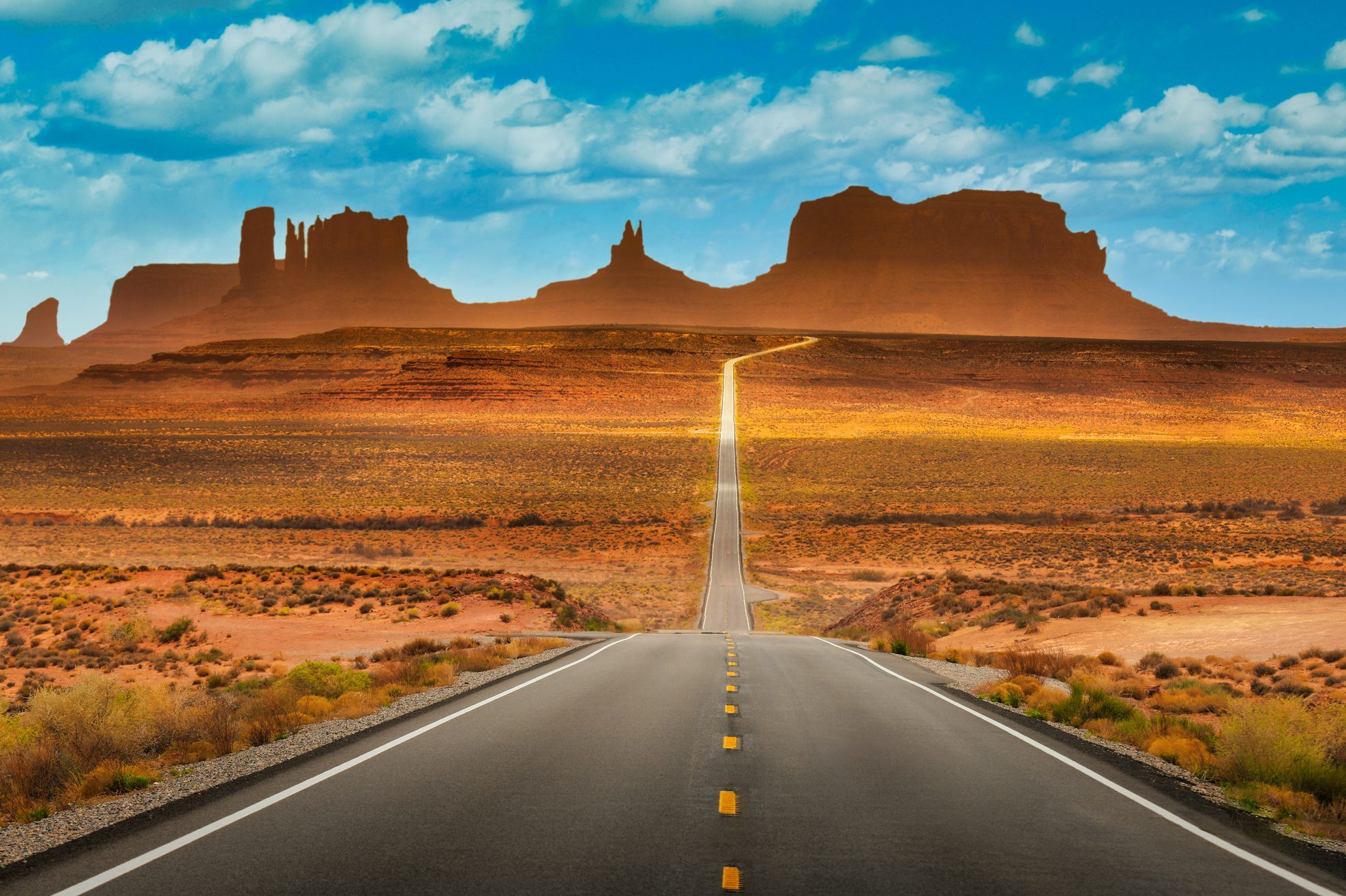 A straight road stretches toward distant mesas under a vibrant blue sky with white clouds, surrounded by desert landscape.