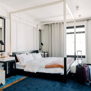 Bed framed by curtains sits next to a side table, across from a suitcase, in a modern hotel room with blue carpeting.