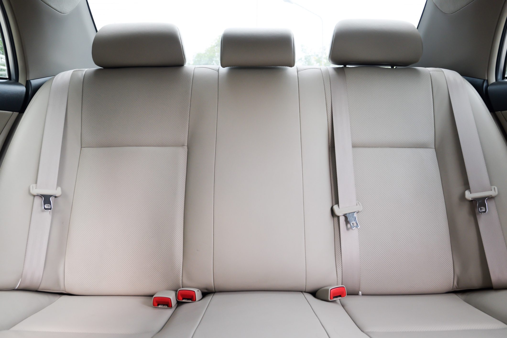 Beige car back seat, with three headrests and two seatbelts fastened. Light enters through the rear windshield, highlighting the upholstery.