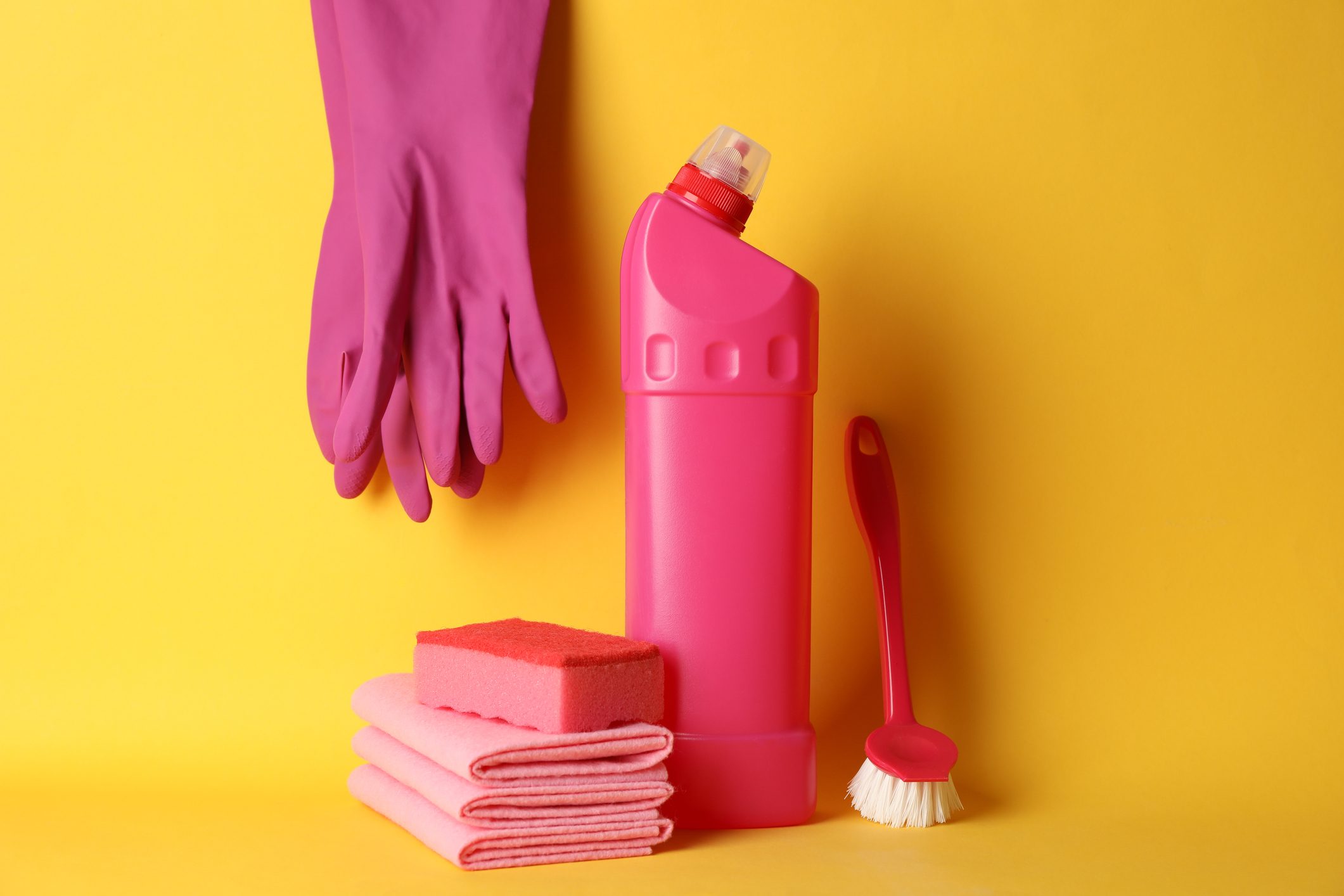 Pink cleaning supplies, including gloves, a bottle, sponge, cloths, and a brush, are arranged together against a bright yellow background.