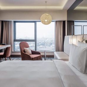 Hotel room features a neatly made bed, desk with chair, and armchair beside large windows overlooking a snowy cityscape, illuminated by warm lighting.