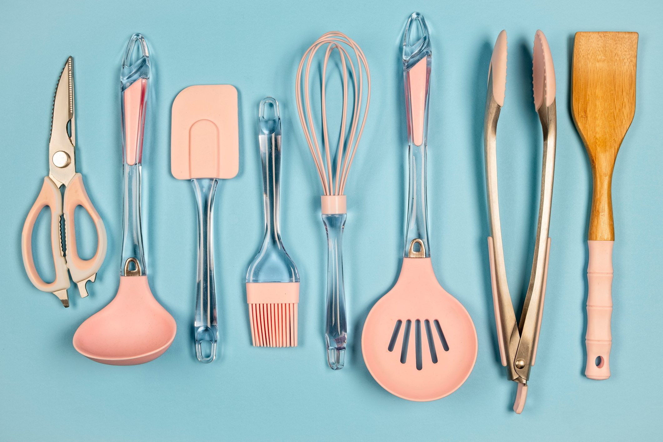 Kitchen utensils arranged side by side on a blue background, including scissors, spatula, whisk, and tongs, all with pink and clear handles.