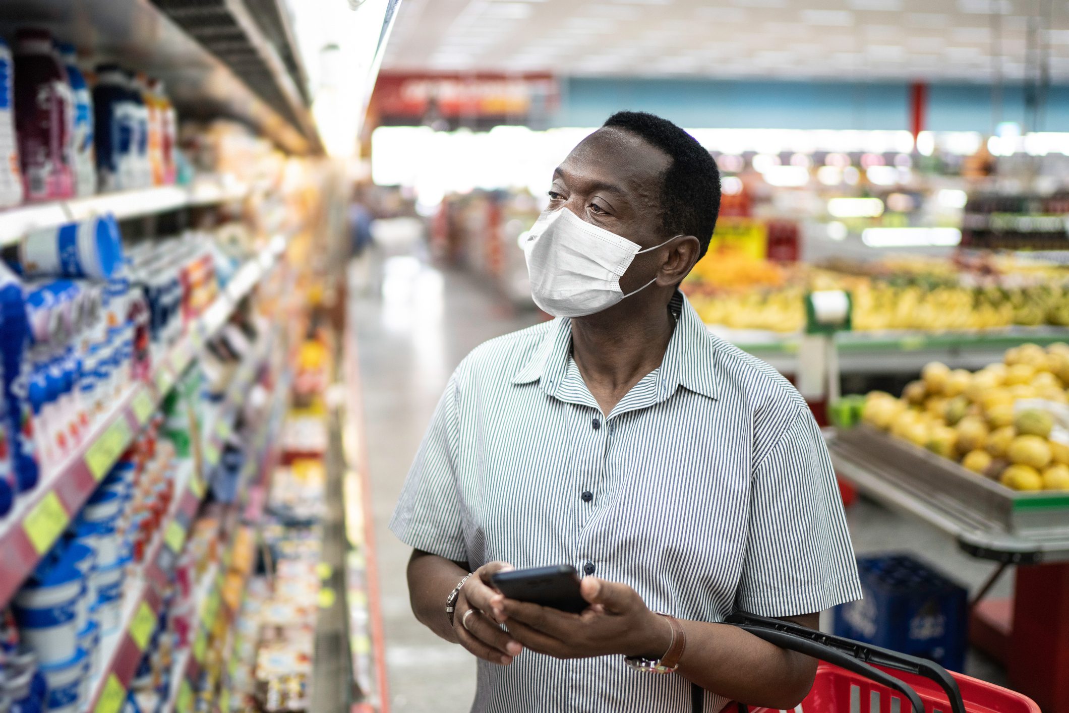 Mature man using mobile and choosing products in supermarket - using face mask