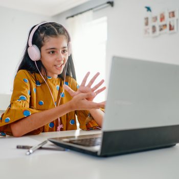 A girl wearing headphones gestures during a video call using a laptop in a brightly lit room.