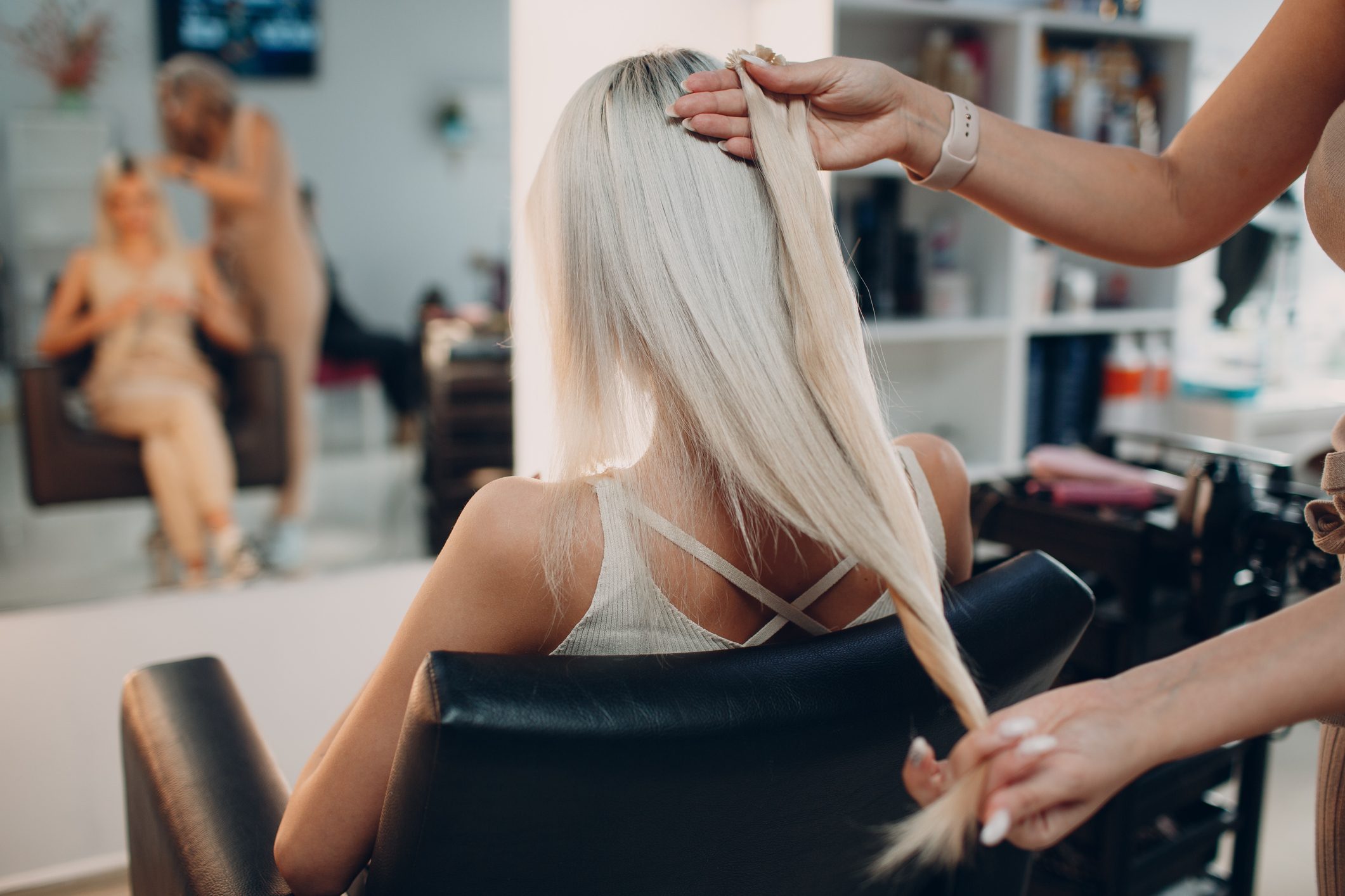 Hair stylist adjusts long blonde hair inside a salon, with a seated client reflected in the mirror.