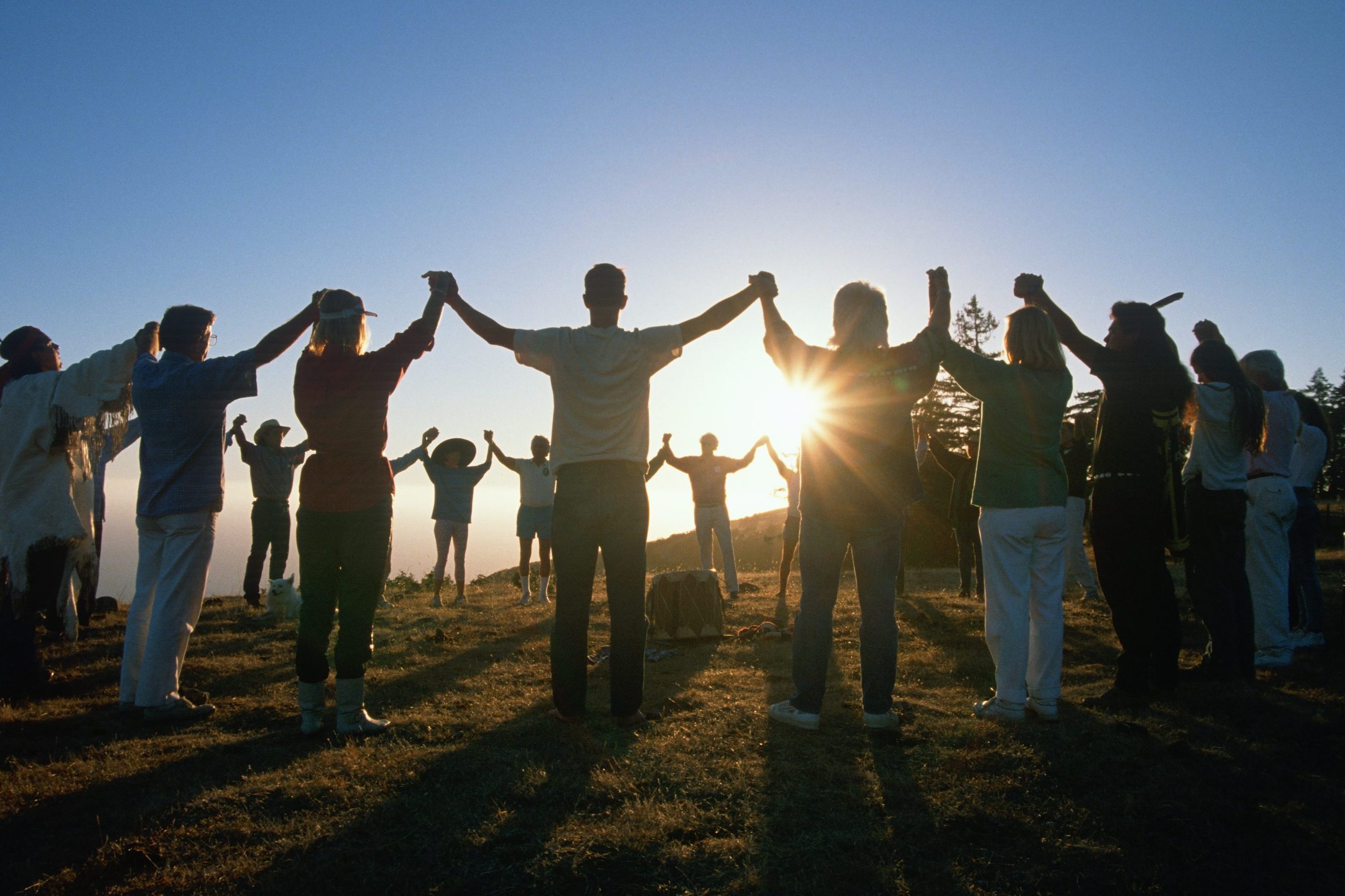 People hold hands, forming a circle against the setting sun on a grassy field. Trees line the background under a clear sky.