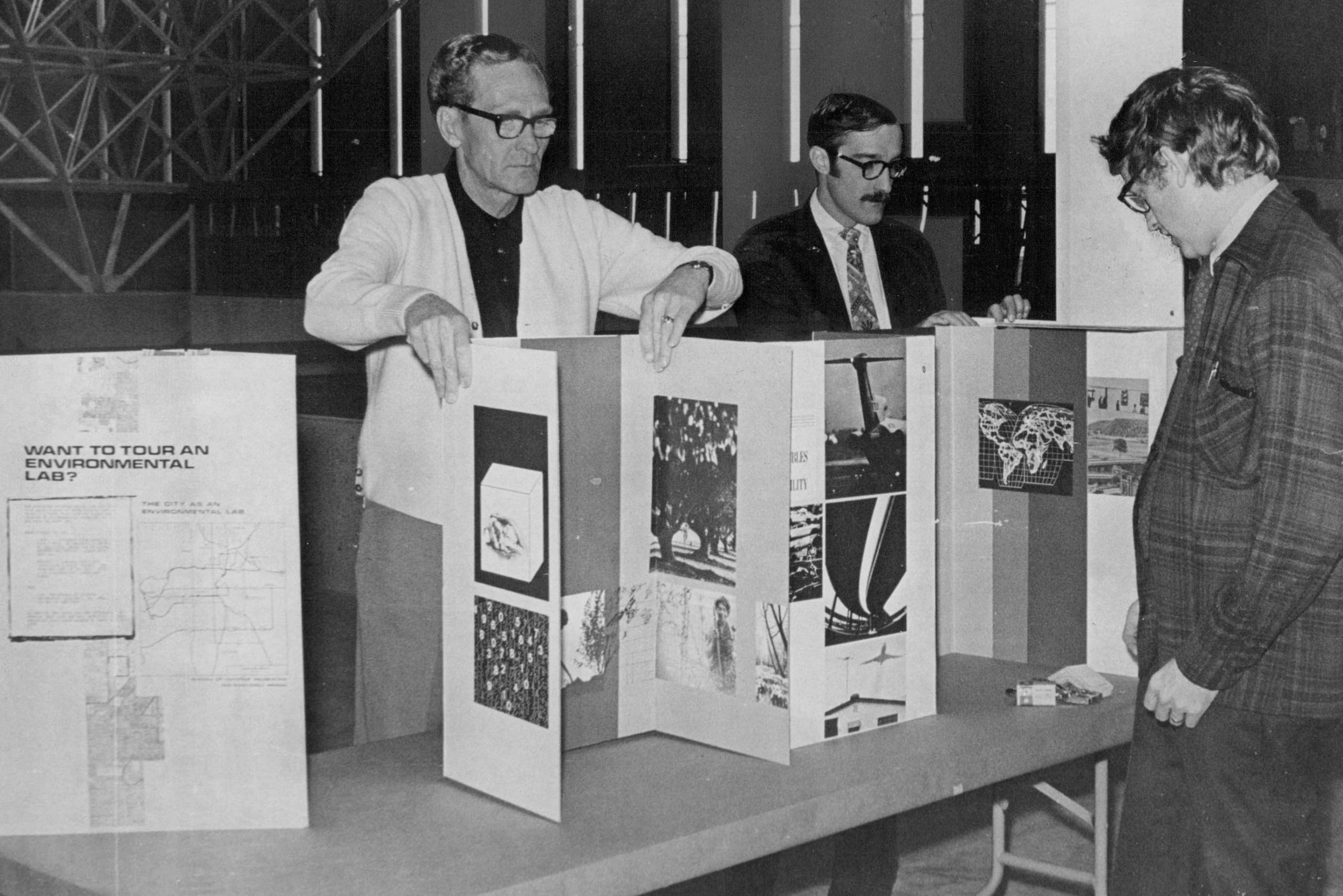 Three men examine a presentation board displaying environmental images and text, including "WANT TO TOUR AN ENVIRONMENTAL LAB?", set indoors.