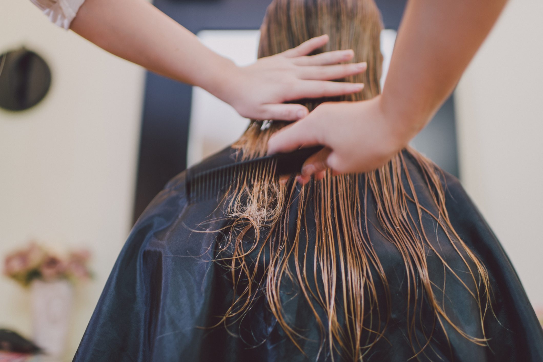 Hands comb wet hair on a seated person, covered by a black cape, in a salon setting.
