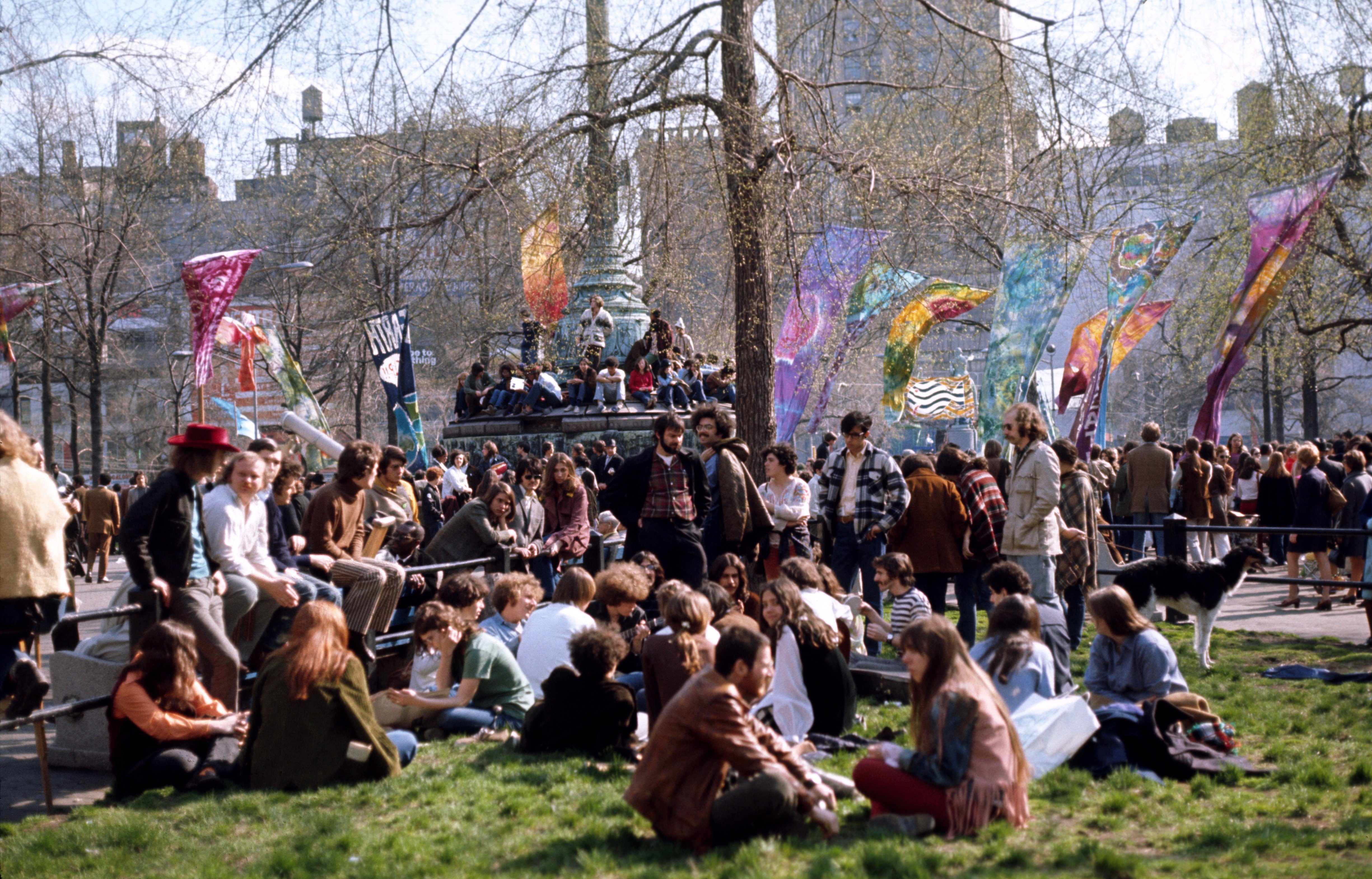 People gather in a park, sitting and standing amidst colorful flags and trees, enjoying a lively outdoor event.