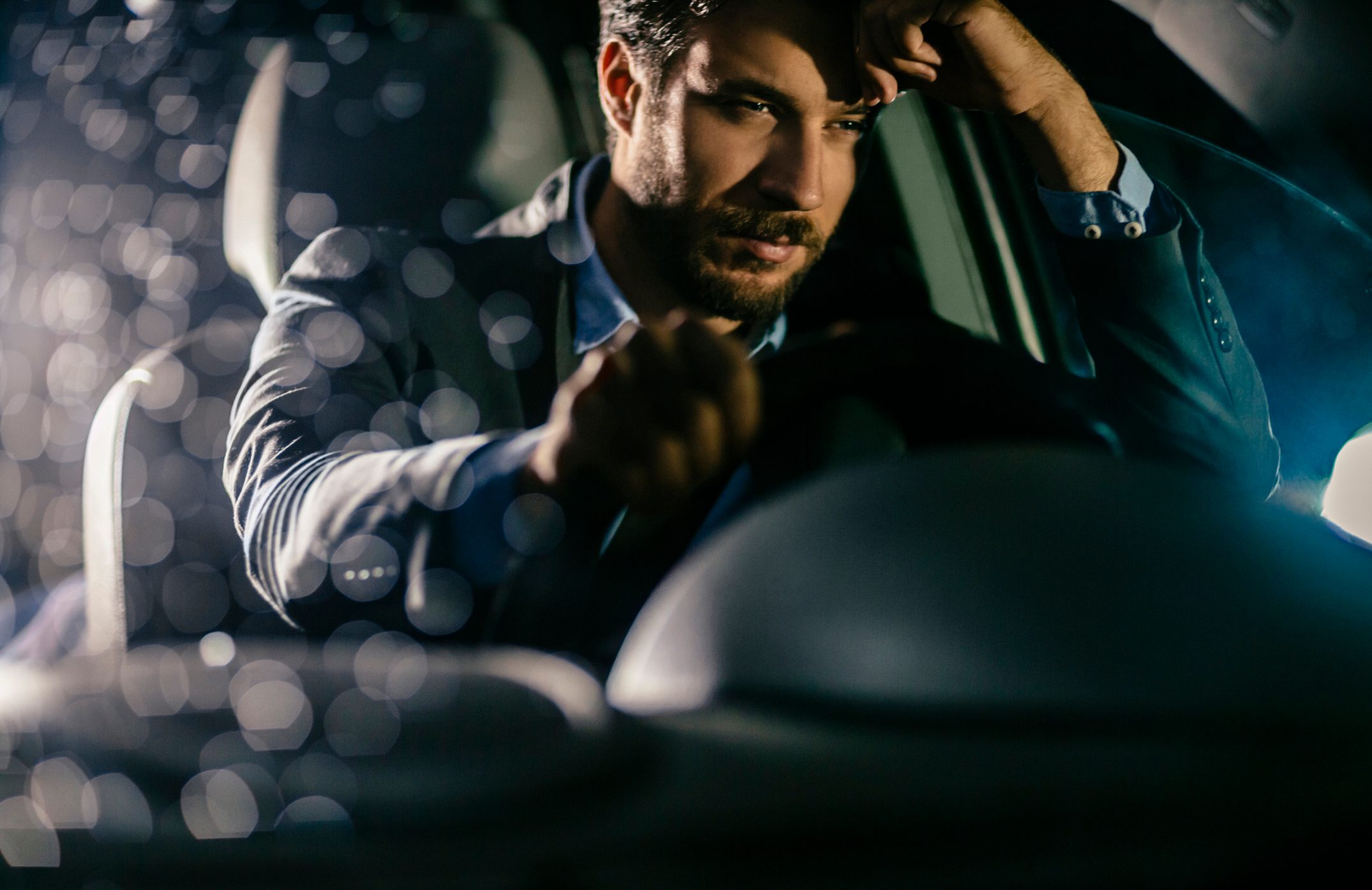 A man sits in a car at night, holding the steering wheel, with a focused expression and blurred lights in the background.