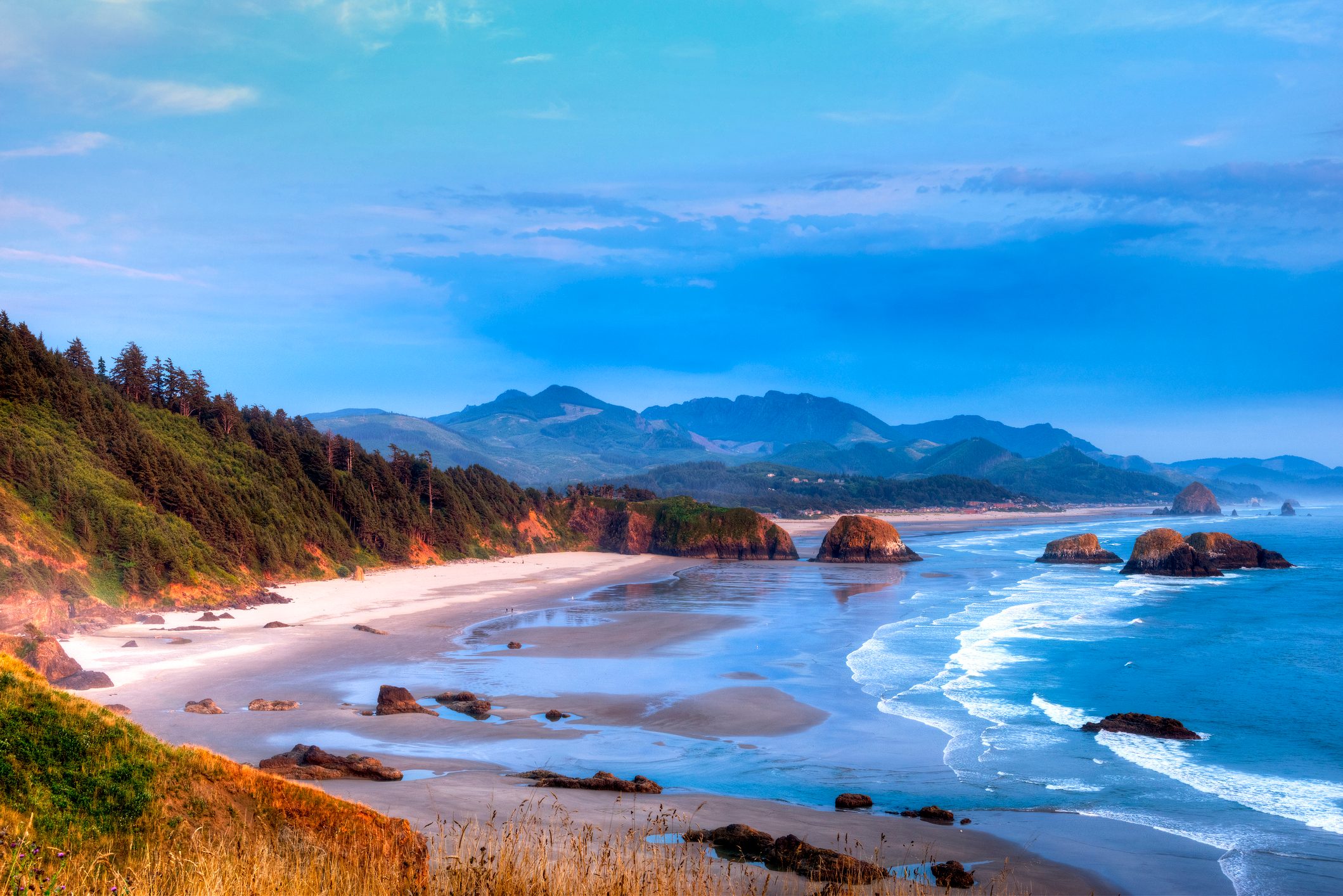 Beach coastline curves alongside serene blue waves, with forested hills and distant mountains under a clear sky.
