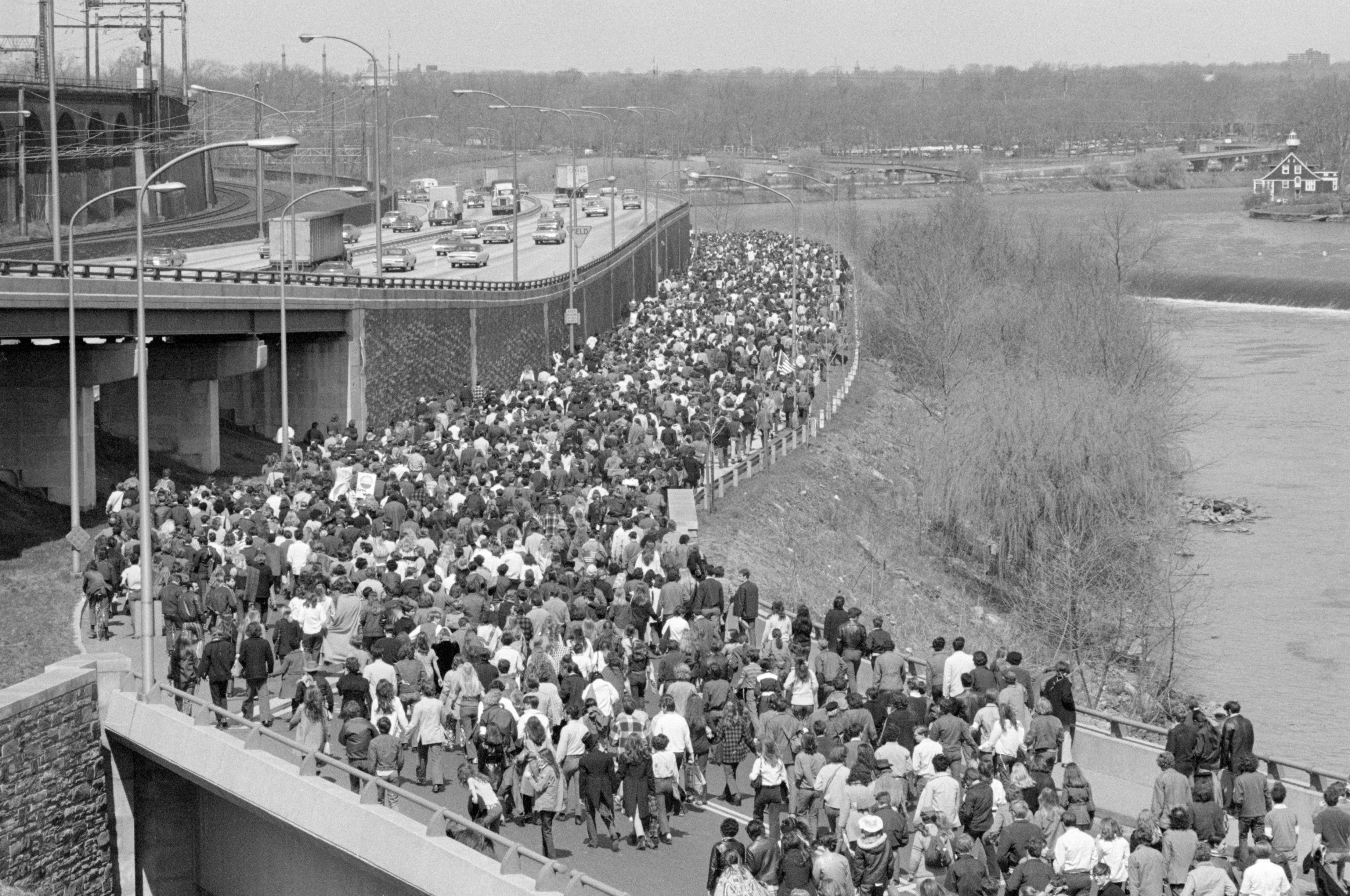 A large crowd walks beside a highway, near a river, with cars on the road and trees lining the background.