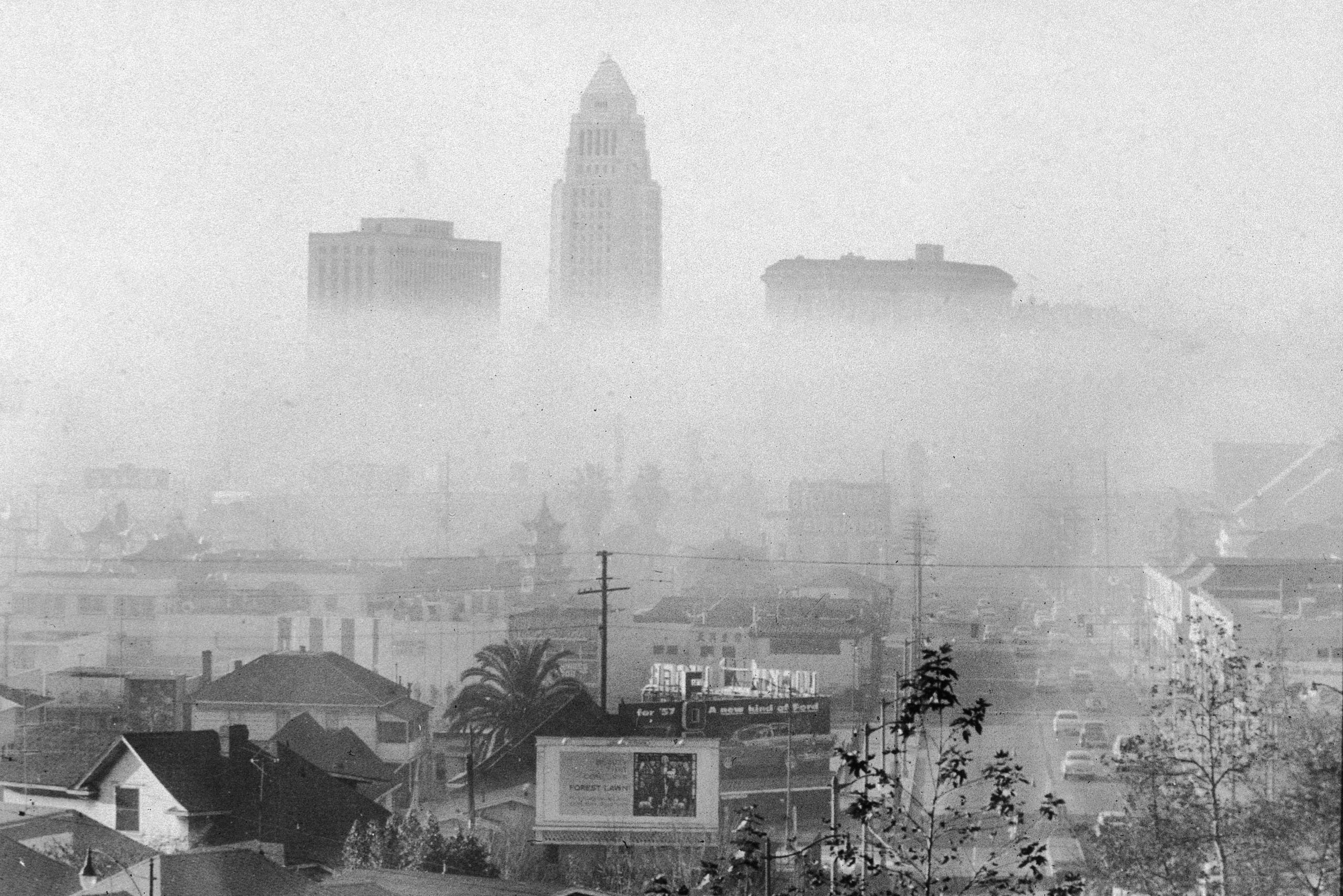 Skyscrapers stand obscured by thick smog, overlooking a busy urban street with cars and billboards.