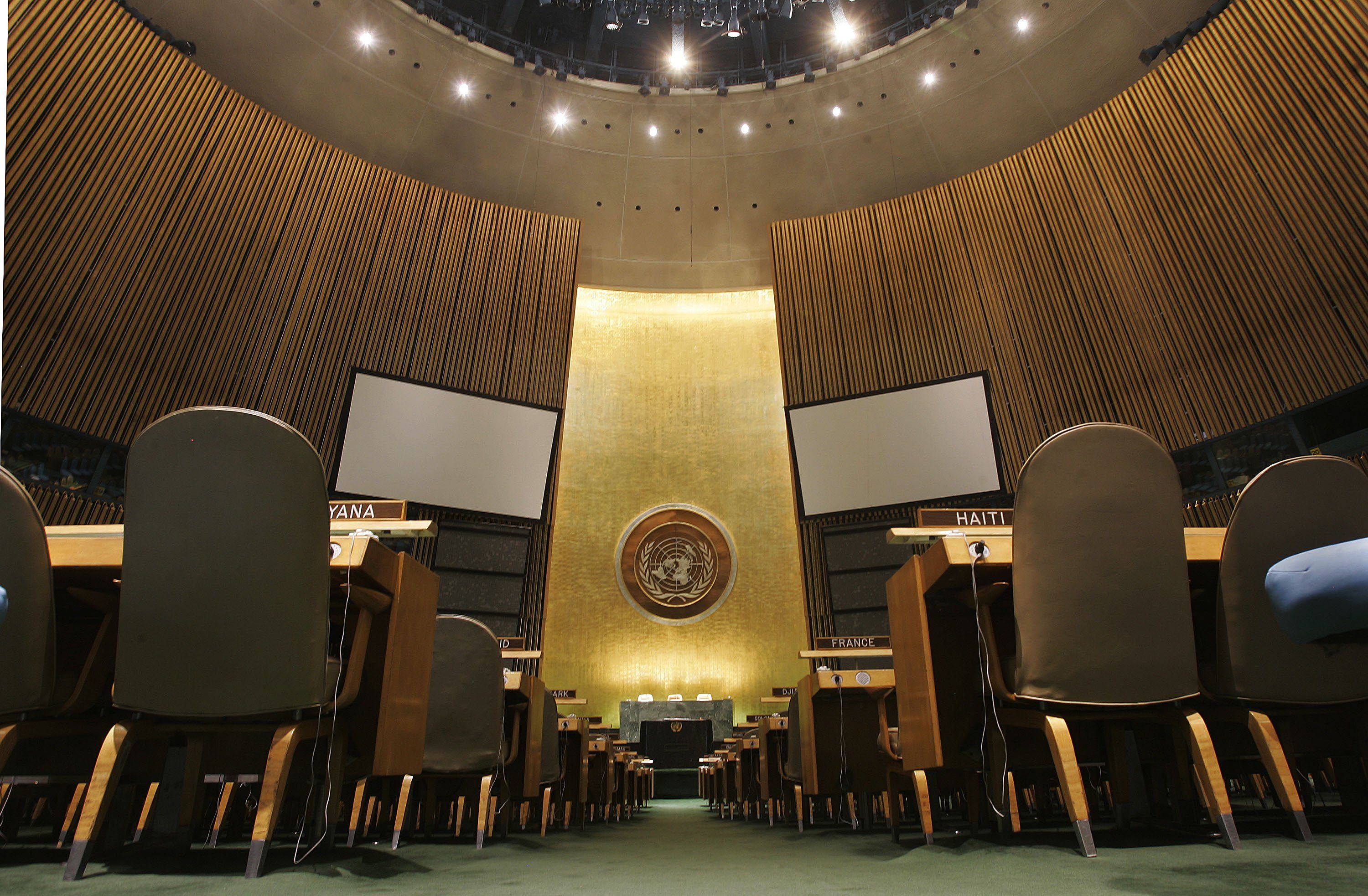 Desks with country nameplates sit empty, facing large screens and a United Nations emblem, within a grand, circular assembly hall.
