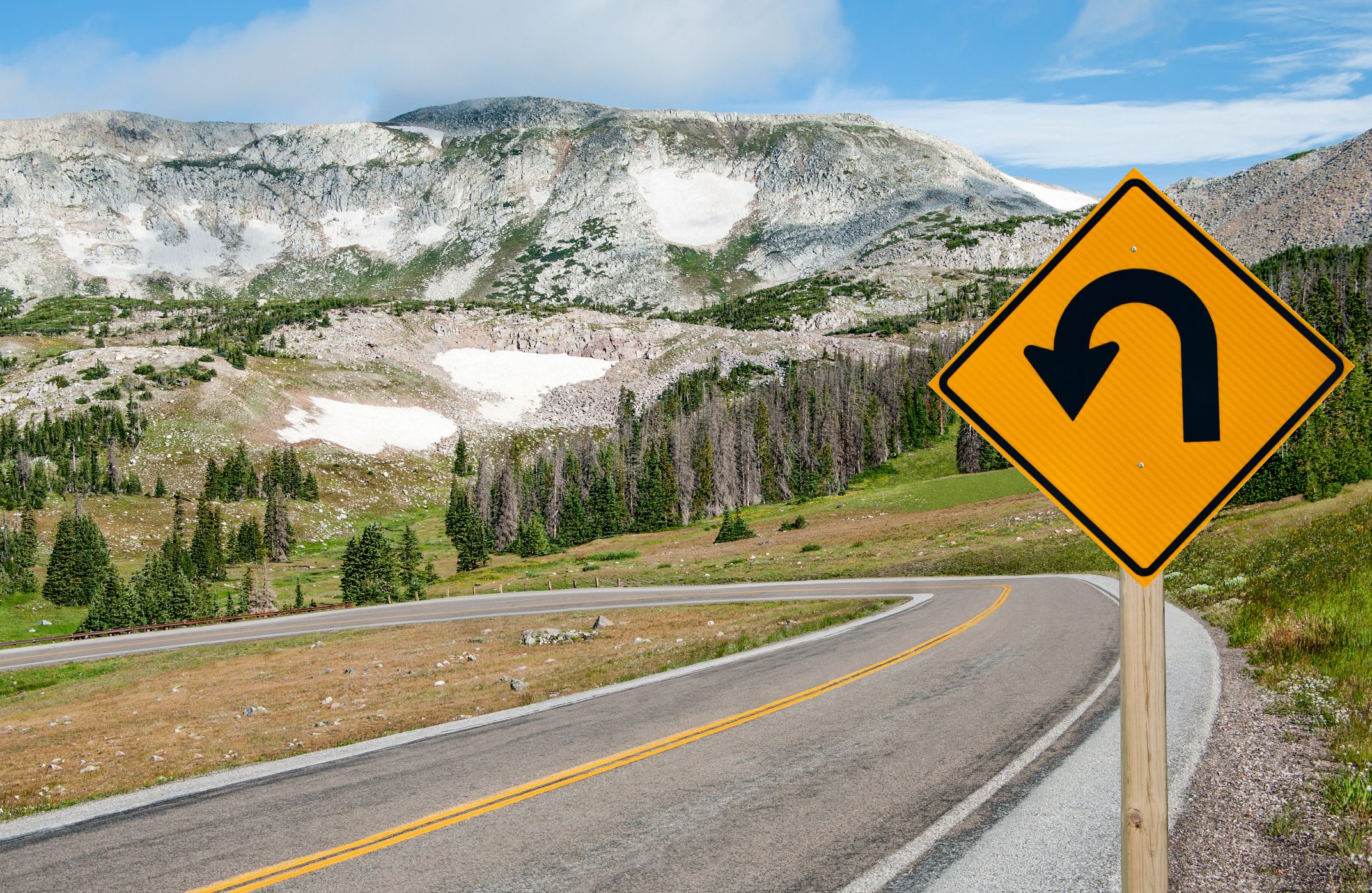 U-turn road sign indicating direction on a winding mountain highway amidst lush green landscape and rocky peaks under a partly cloudy sky.