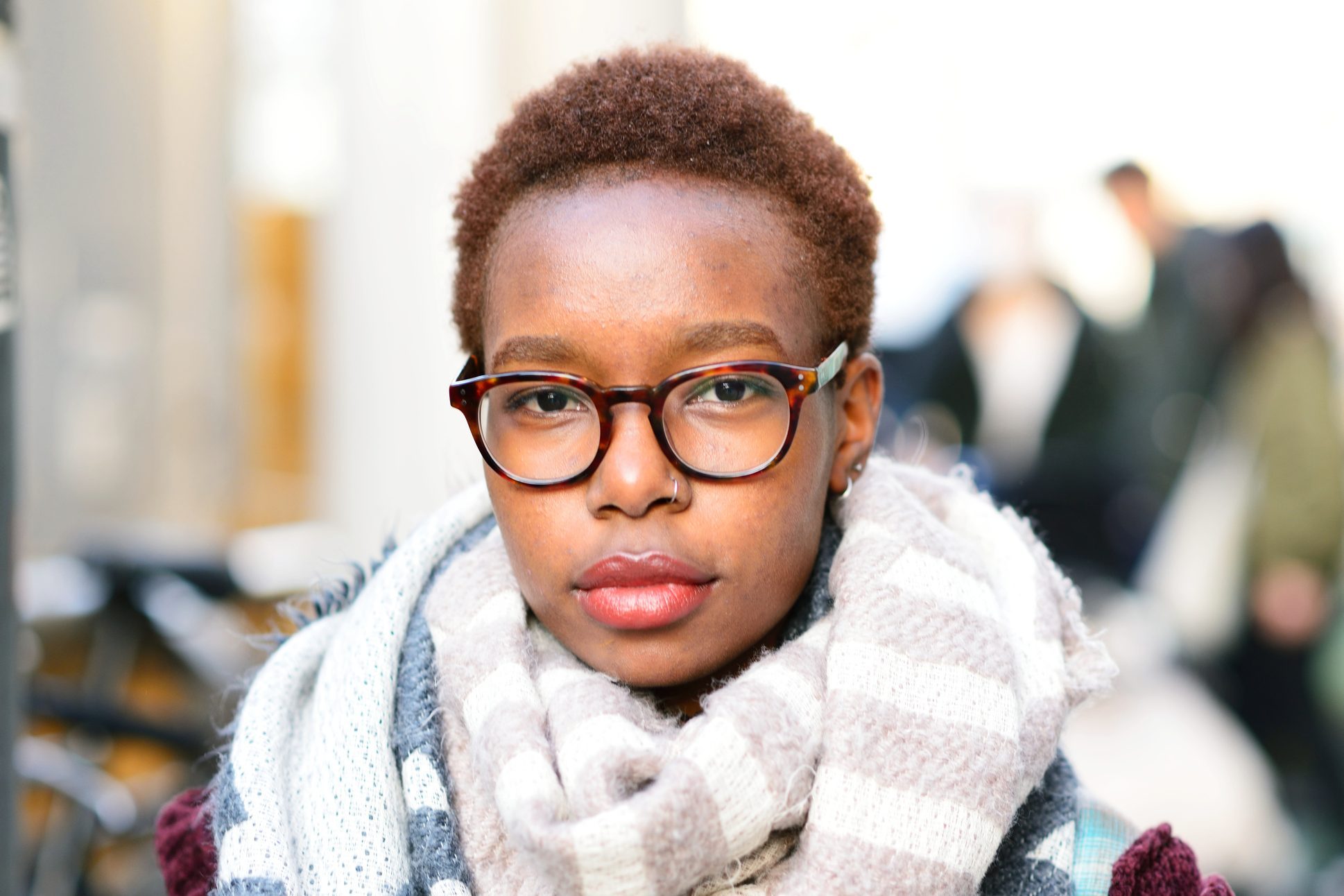 Person wearing glasses looks directly ahead, wrapped in a striped scarf. The background is blurred, suggesting an outdoor urban setting.
