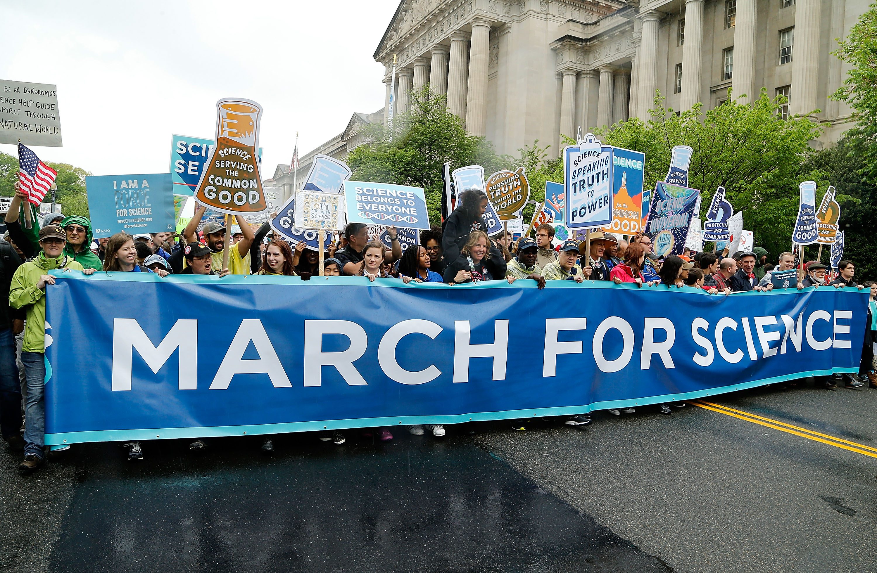 Protesters hold signs behind a "MARCH FOR SCIENCE" banner, demonstrating on a city street with a historical building and greenery in the background.