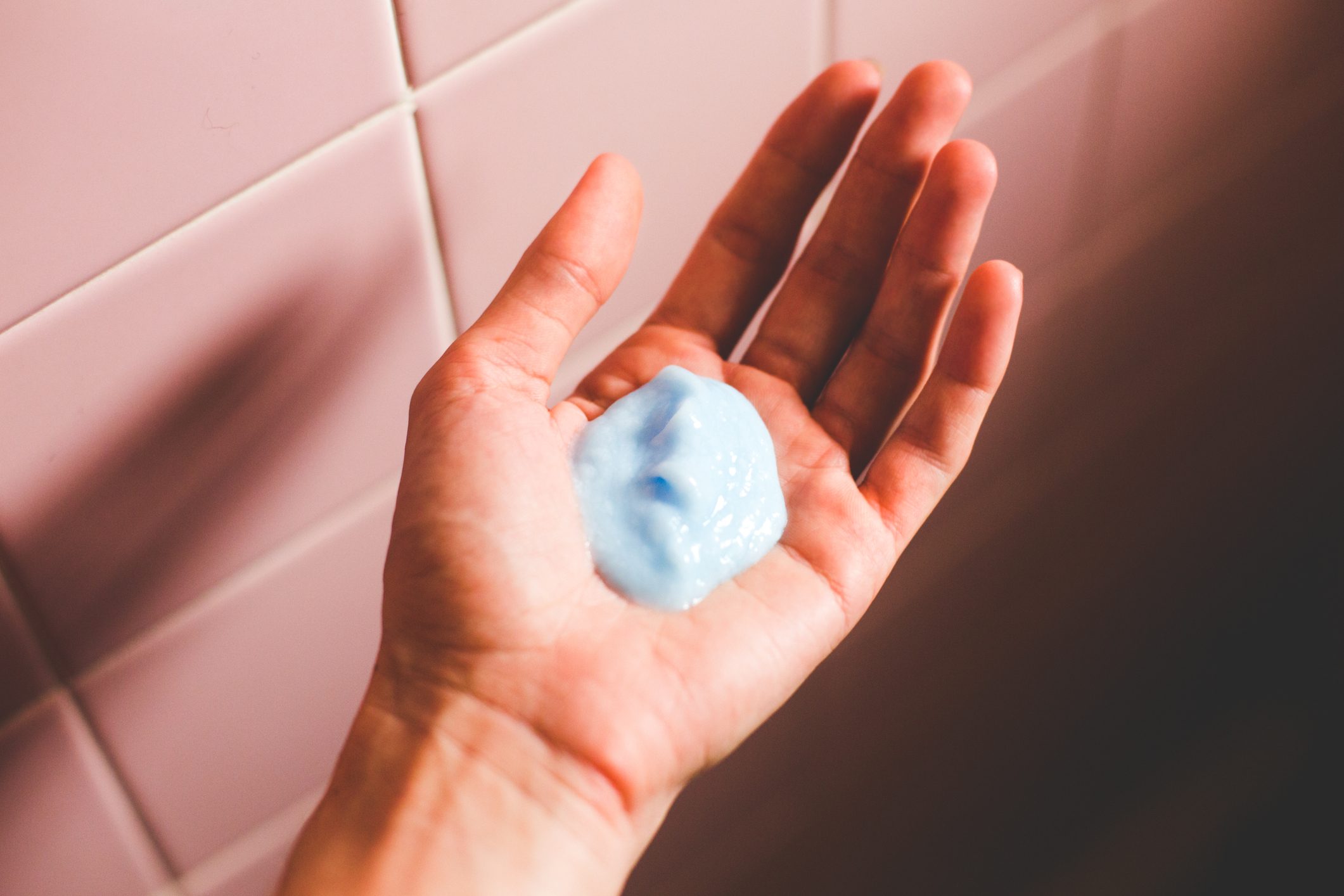 A hand holds blue gel amidst pink tiled walls, possibly in a bathroom setting.