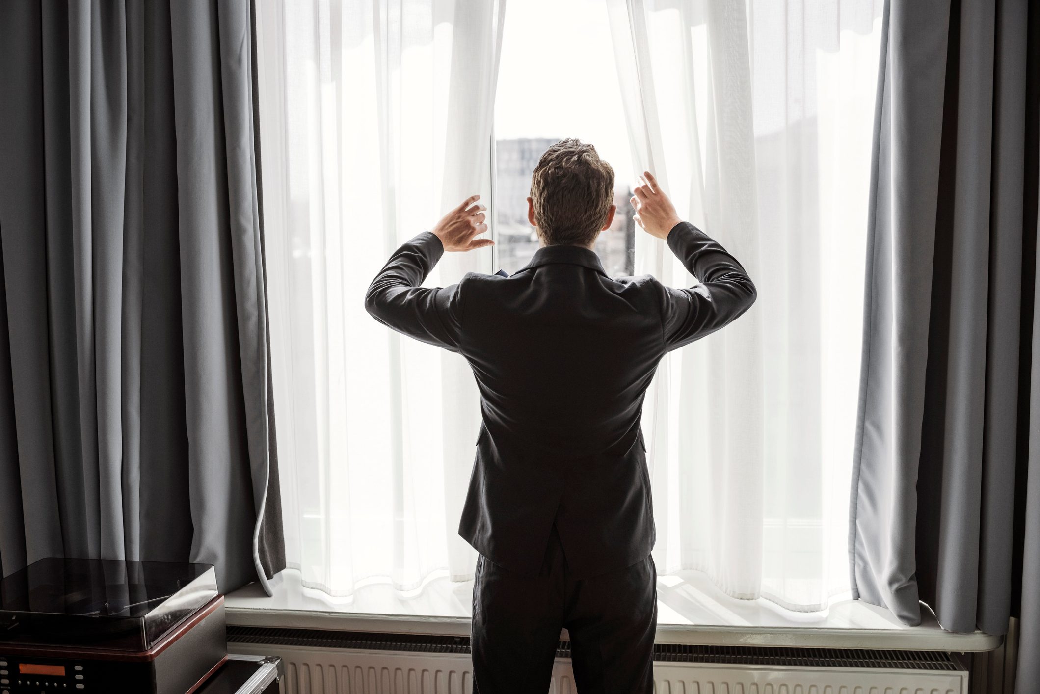 Rear view of businessman opening white curtain on window in hotel room