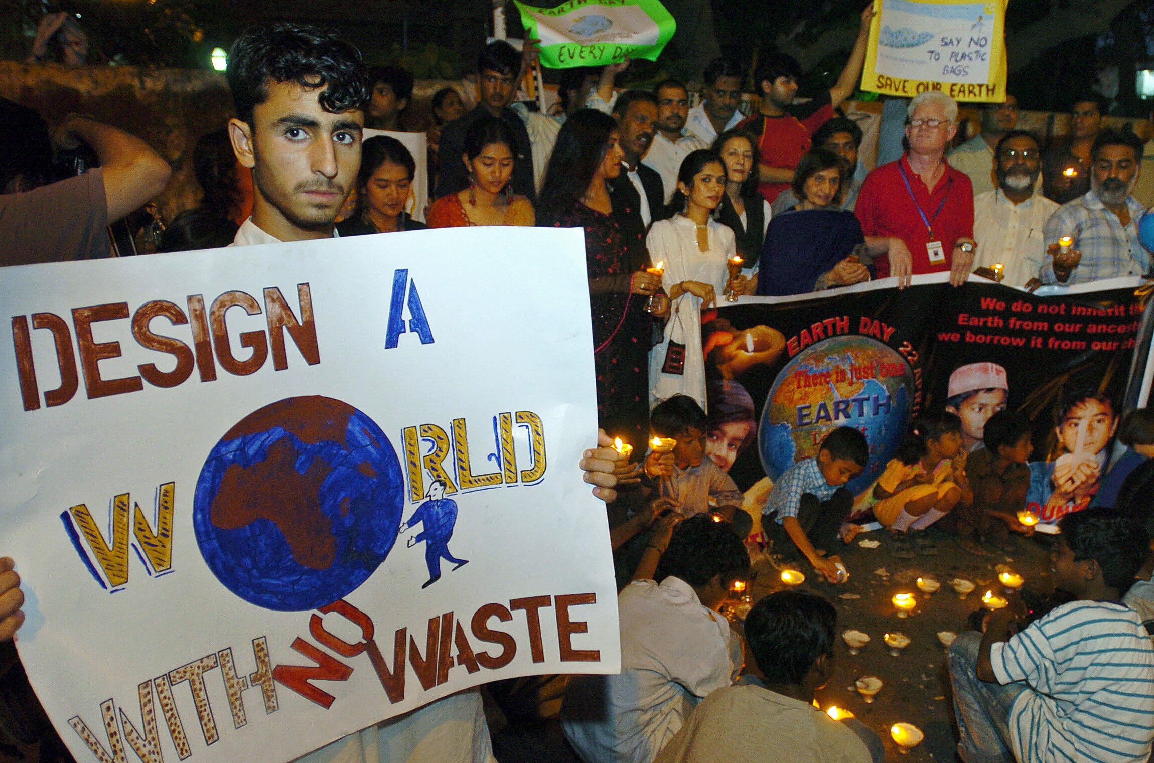 People hold candles and protest signs, like "DESIGN A WORLD WITH NO WASTE," gathering for an environmental demonstration outdoors at night.
