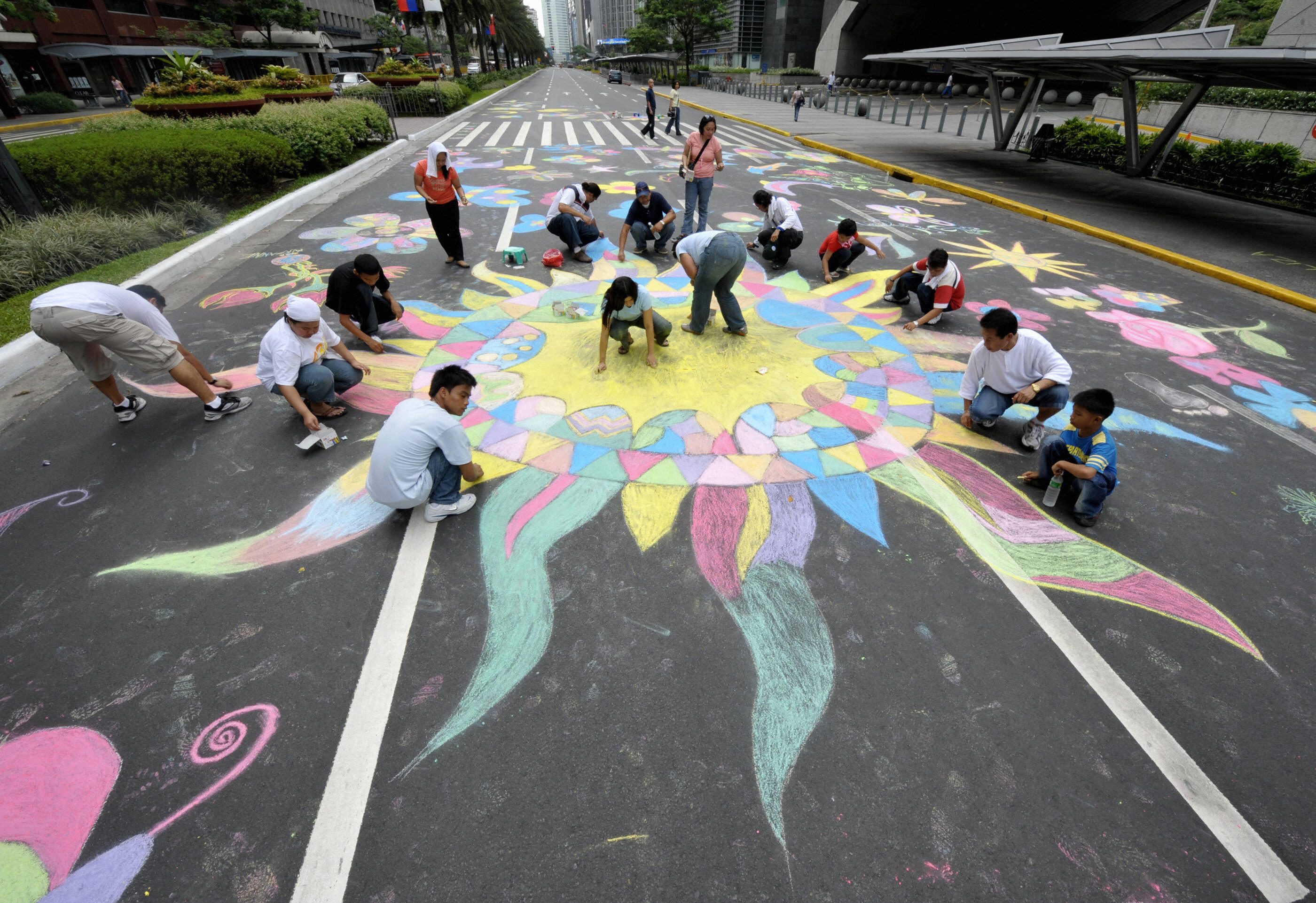 People create colorful chalk designs on a city street, surrounded by buildings.