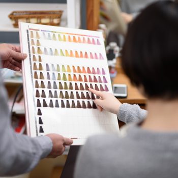 A hair color chart is held while someone points at a color, inside a salon setting.
