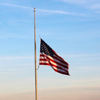 Flag flies at half-mast on a flagpole, against a clear blue sky background.