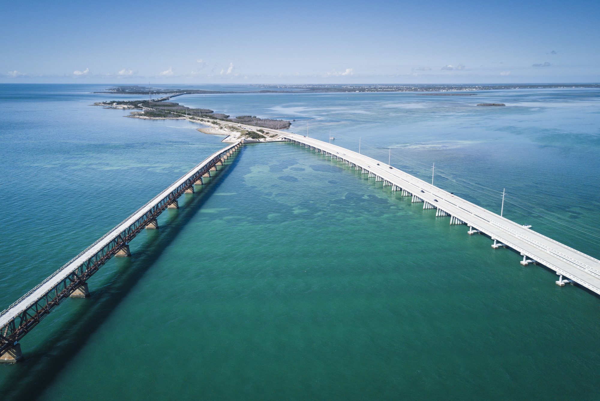 Two parallel bridges span over turquoise water, connecting distant landmasses, under a clear blue sky.